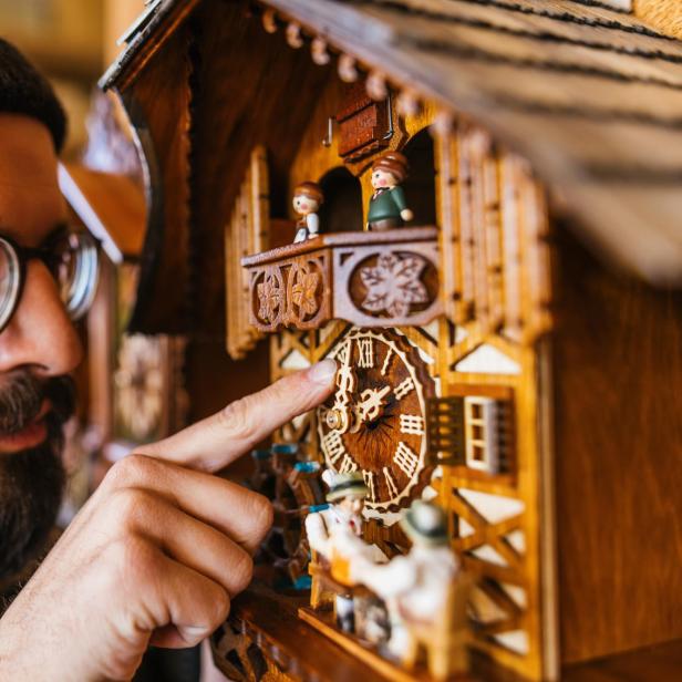 Ein Mann mit Brille und Mütze stellt die Zeit an einer Kuckucksuhr.