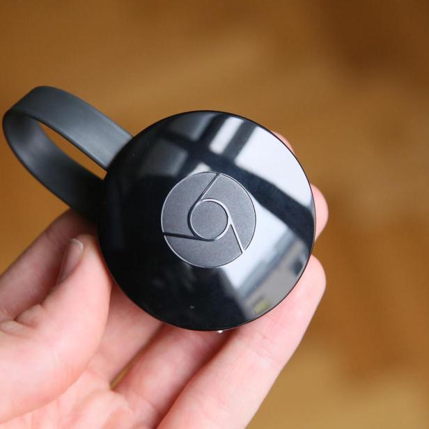 Eine Hand hält einen schwarzen Google Chromecast vor unscharfem Hintergrund.