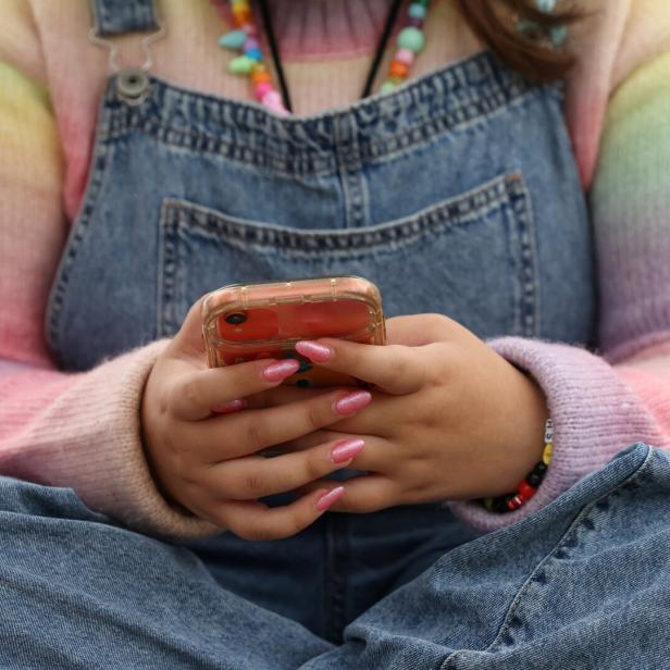 Person in Latzhose hält ein Smartphone mit pink lackierten Nägeln.