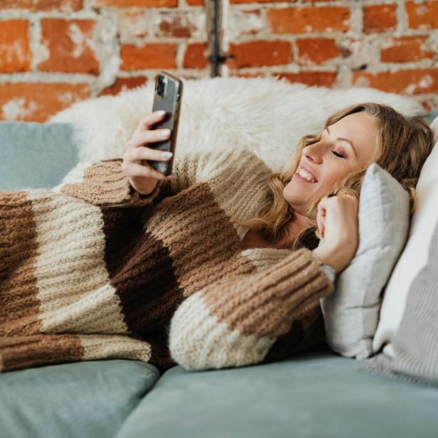 Eine Frau in einem braun-gestreiften Pullover liegt auf der Couch und blickt auf ihr Smartphone