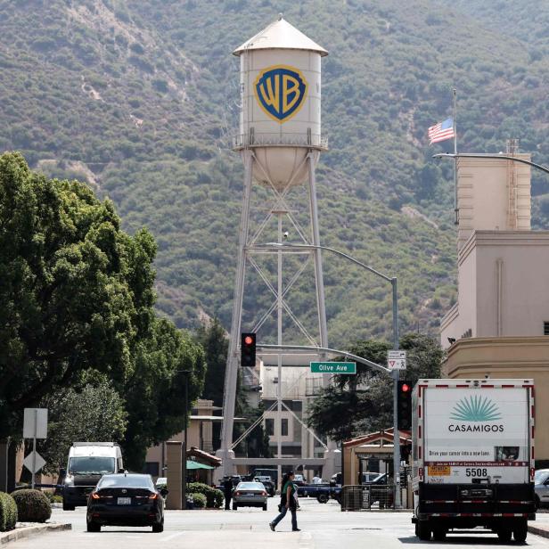 Großer Wasserturm mit WB-Logo vor bewaldetem Hügel, Autos und Fußgänger an einer Kreuzung.