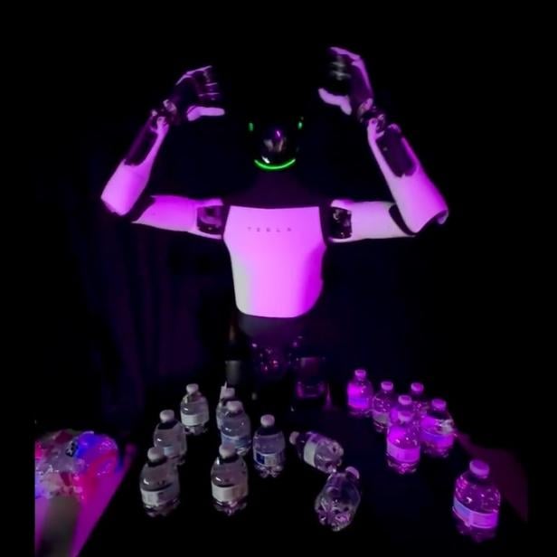 Der Tesla-Roboter Optimus steht auf einem Event in Miami hinter einem Tisch mit mehreren Wasserflaschen und hebt beide Arme.