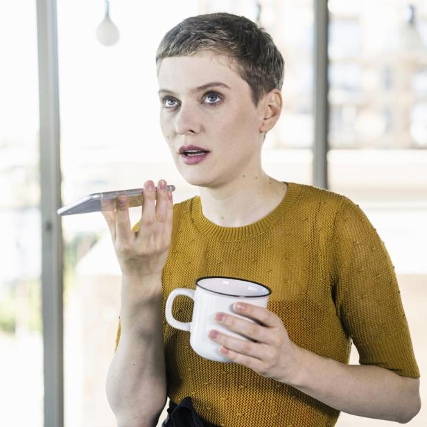 Eine Frau mit kurzem Haar telefoniert mit dem Handy und hält eine Tasse.