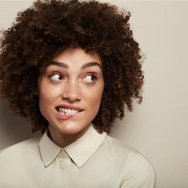 Eine junge Frau mit lockigem Haar schaut zur Seite und beißt sich nachdenklich auf die Lippe, passend zum Thema Zahnpflege.