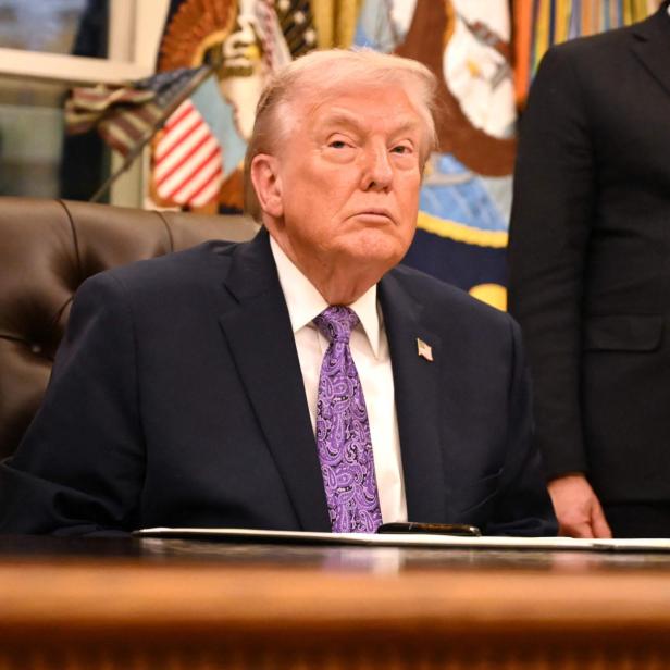 Donald Trump sitzt an einem Schreibtisch, trägt einen dunklen Anzug und eine lila Krawatte.