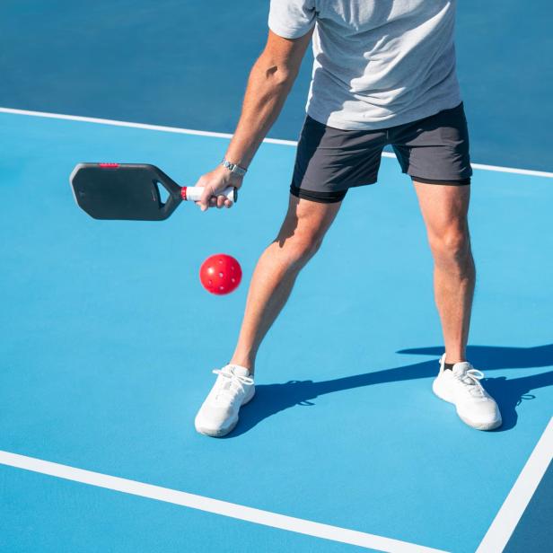 Person spielt Pickleball mit schwarzem Schläger und rotem Ball auf blauem Spielfeld.