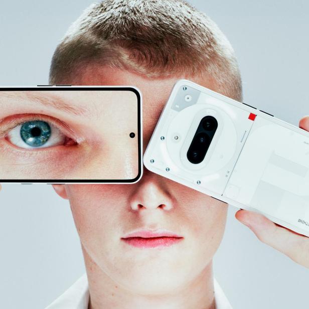 Eine Person hält ein Smartphone mit einem vergrößerten Auge auf dem Display vor ihr Gesicht und daneben ein weiteres Smartphone mit sichtbarer Rückseite.
