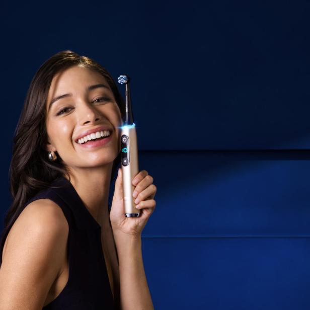 Eine Frau hält lächelnd die Oral-B iO10 Gold Special Edition Zahnbürste vor einem dunkelblauen Hintergrund.