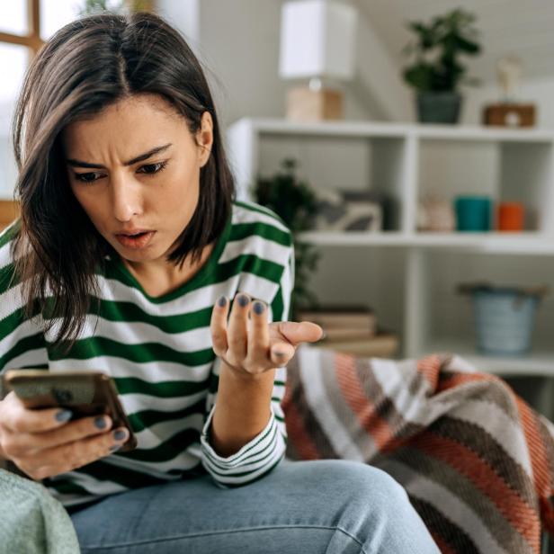 Eine junge Frau mit grün-weiß gestreiftem Pullover schaut verärgert auf ihr Smartphone und gestikuliert mit der Hand.