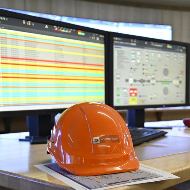 Ein orangefarbener Schutzhelm liegt auf einem Schreibtisch vor mehreren Monitoren mit technischen Übersichten und Tabellen.