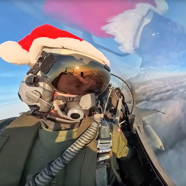 Pilot mit Weihnachtsmütze im Cockpit, dahinter Kampfjets in Weihnachtsbaum-Formation am Himmel.