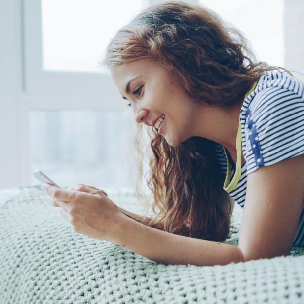 Junge Frau liegt lächelnd auf dem Bett und nutzt ihr Smartphone vor einem großen Fenster.