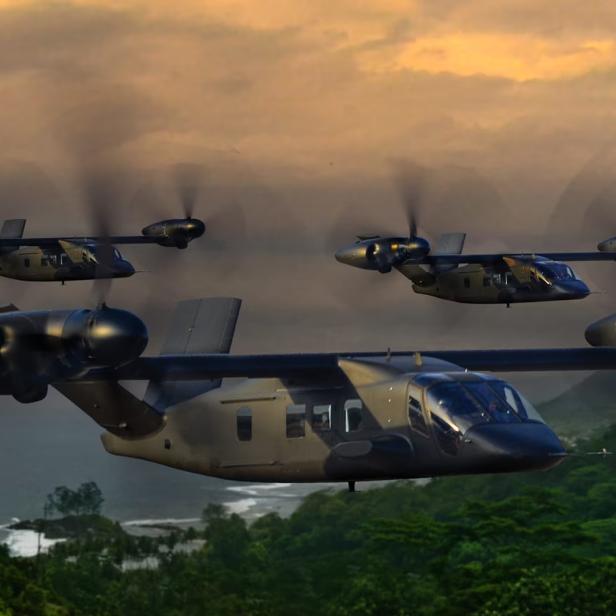 Vier militärische Kipprotorflugzeuge fliegen bei Sonnenuntergang über eine grüne, bewaldete Küstenlandschaft.