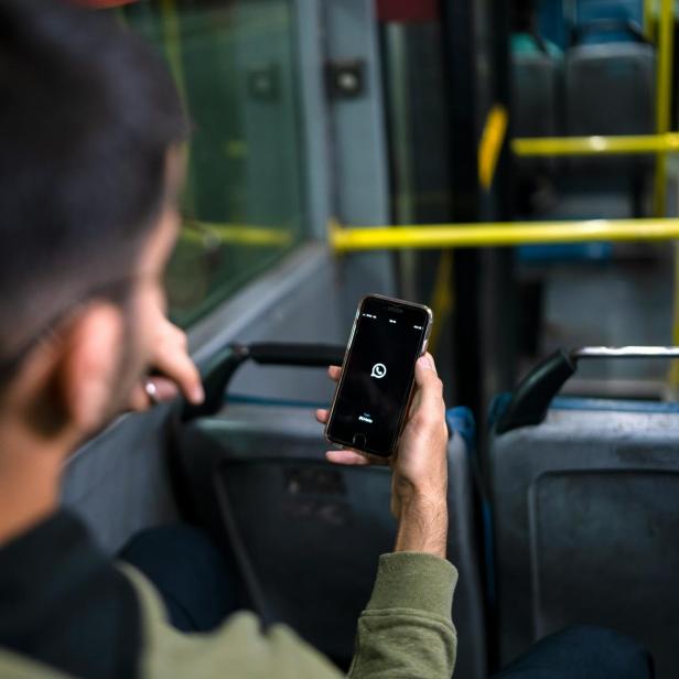 Ein Mann sitzt in öffentlichem Verkehrsmittel mit Smartphone, auf dem WhatsApp geöffnet wird 