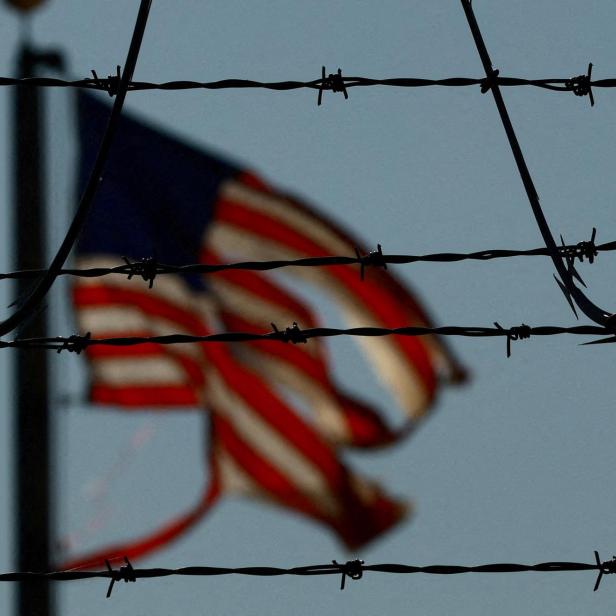 Stacheldraht im Vordergrund, im Hintergrund weht unscharf eine amerikanische Flagge im Wind.