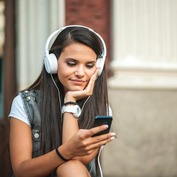 Junge Frau mit langen dunklen Haaren hört mit weißen Kopfhörern Musik und schaut auf ihr Smartphone.