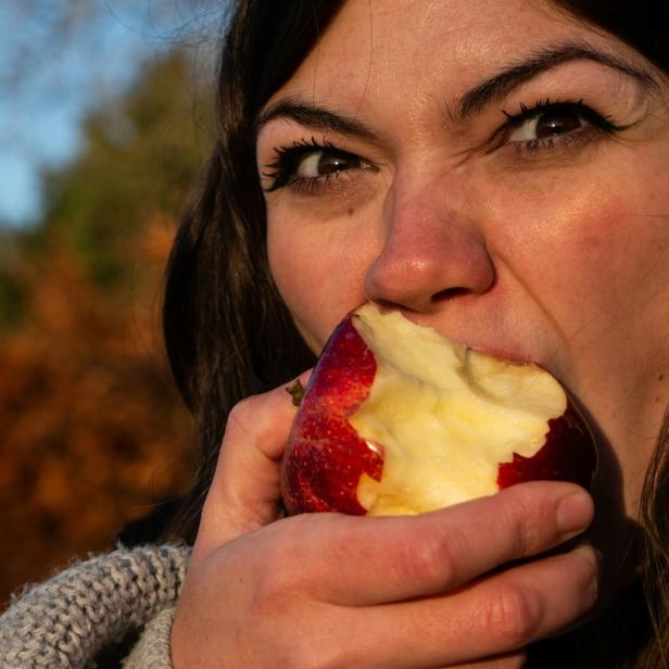 Frau beißt in einen roten Apfel im Freien mit herbstlichem Hintergrund.