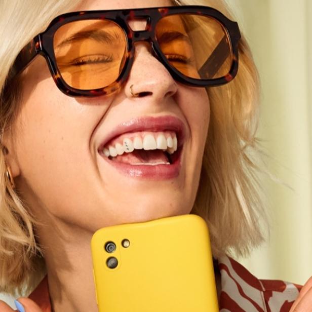 Lächelnde Frau mit großer Sonnenbrille hält ein gelbes Smartphone in der Hand.