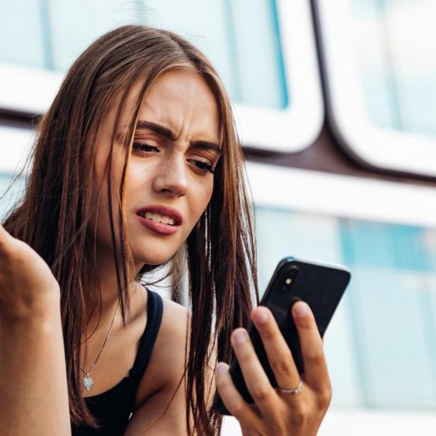 Junge Frau schaut mit verwirrtem Gesichtsausdruck auf ihr Smartphone und hebt fragend die Hand.
