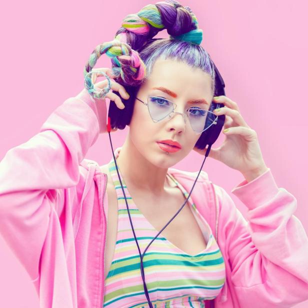 Junge Frau mit buntem Zopf, Kopfhörern und modischer Brille trägt gestreiftes Top und pinke Jacke vor rosa Hintergrund.