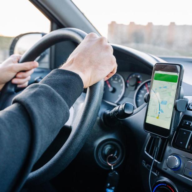 Eine Person fährt ein Auto und nutzt ein Smartphone mit Navigations-App an der Armatur.
