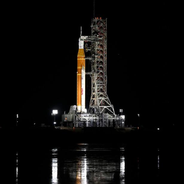 Eine große Rakete steht nachts beleuchtet auf einer Startrampe, umgeben von Scheinwerfern und Gerüsten.