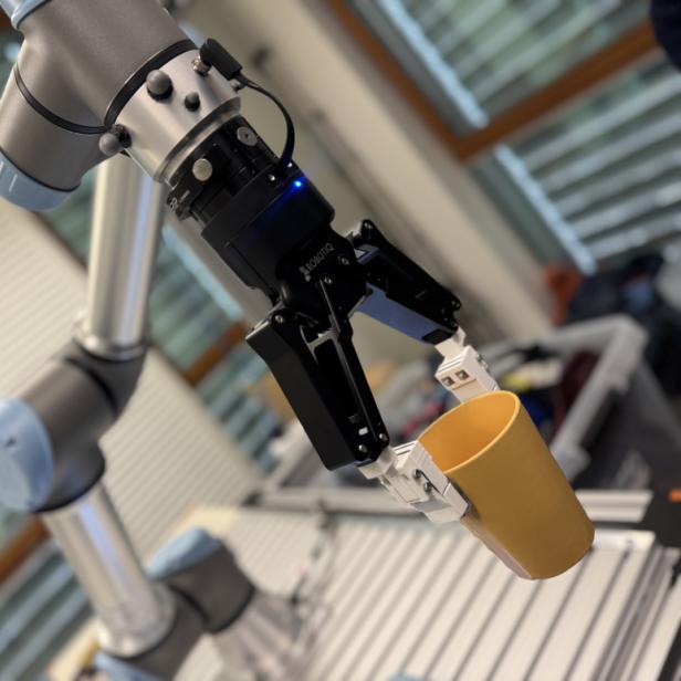Roboterarm mit Greifhand hält einen gelben Becher in einem Laborraum.