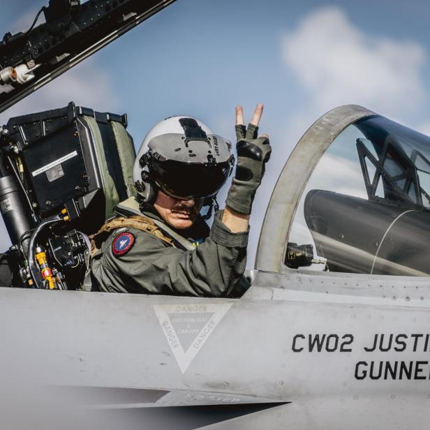Pilot mit Helm und Handschuhen zeigt Victory-Zeichen im Cockpit eines Kampfjets bei offenem Dach.