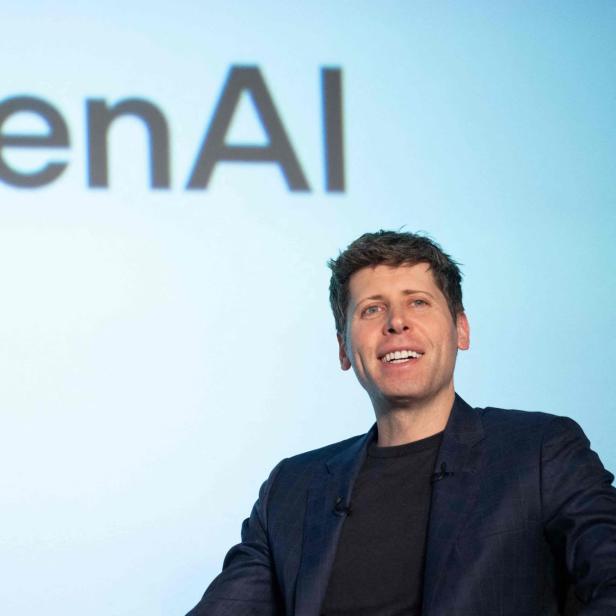 Ein Mann lächelt vor dem "OpenAI"-Schriftzug auf hellblauem Hintergrund.