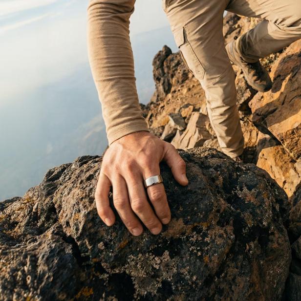 ein Mann in beigem Outfit klettert auf einen Berg, an der Hand hat er einen silberenen Ring