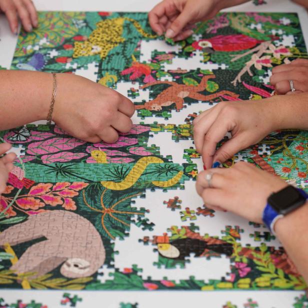 Mehrere Hände setzen gemeinsam ein buntes Puzzle mit Dschungeltieren und Pflanzen zusammen.