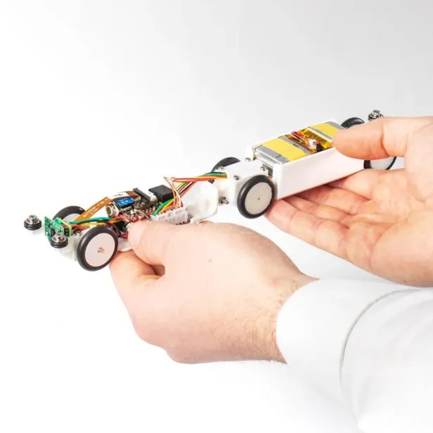 2 Hände halten ein kleines, selbstgebautes Roboterfahrzeug mit sichtbaren elektronischen Bauteilen und Kabeln.