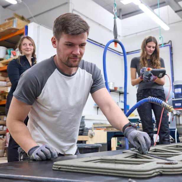 Drei Personen arbeiten mit einem 3D-Betondrucker an einem Fachwerkträger in einer Werkstatt.