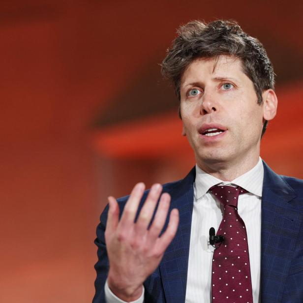 Sam Altman mit Anzug und Krawatte gestikuliert vor einem orangefarbenen Hintergrund.