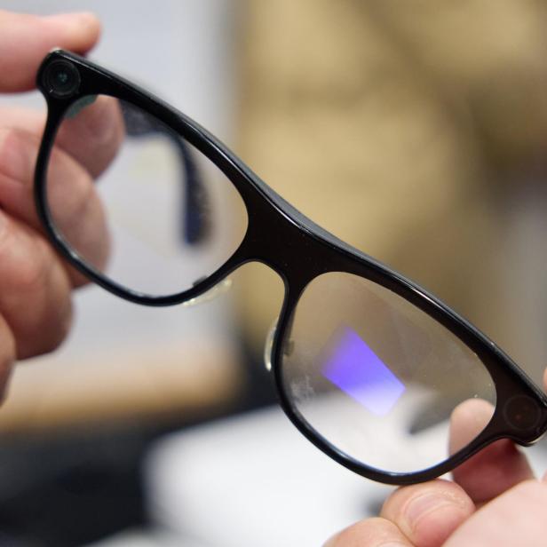 Eine schwarze Brille mit integrierten Kameras an den Ecken wird von zwei Händen gehalten.