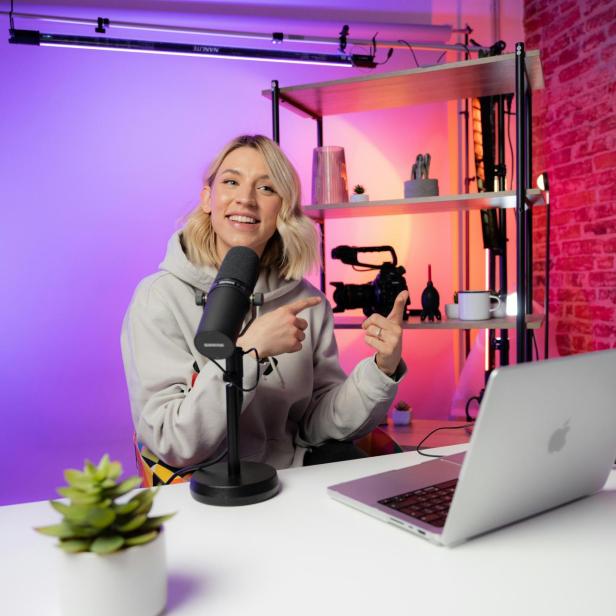 Eine Creatorin sitzt vor Mikrofon und Laptop in einem modernen Studio und spricht, im Hintergrund Kamera-Equipment und bunte Beleuchtung.