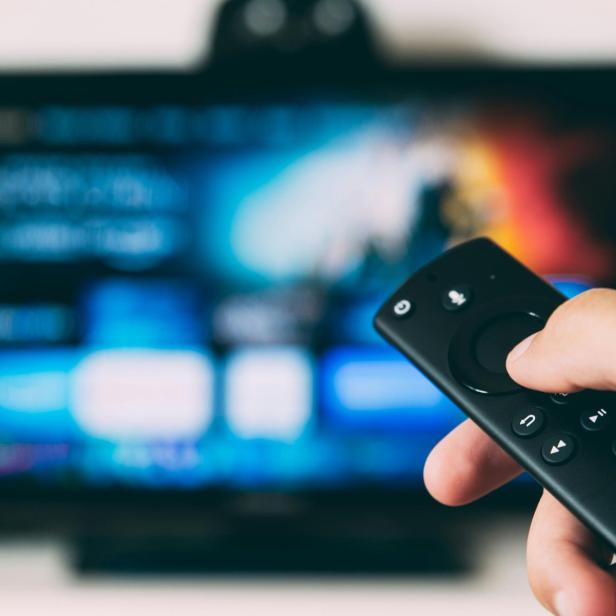 Hand hält Fernbedienung vor unscharfem Fernsehbildschirm mit Streaming-Menü.