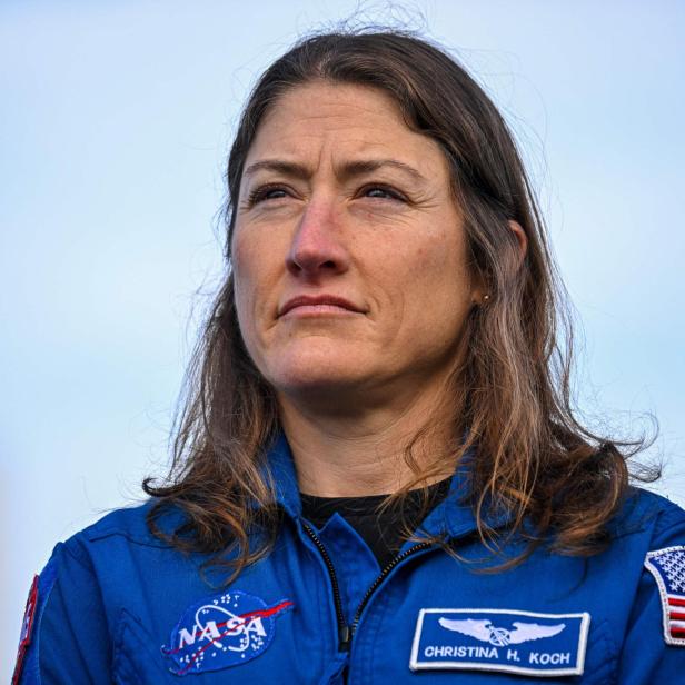 Eine Frau mit langen braunen Haaren trägt einen blauen Raumanzug mit NASA-Logo und US-Flagge vor blauem Himmel.