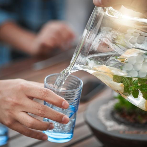 Wasser mit Zitronenscheiben, Minze und Eiswürfeln wird aus einer Glaskaraffe in ein Trinkglas gegossen, um auf die Bedeutung von Wasserqualität im Alltag hinzuweisen.