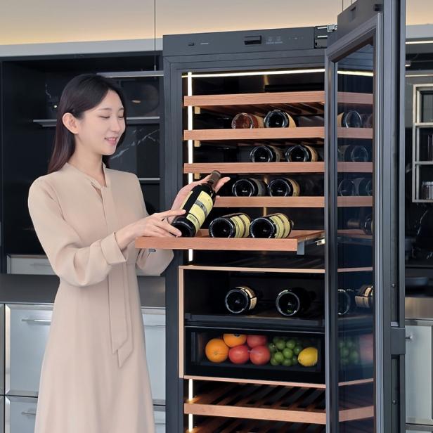 Eine Frau stellt eine Weinflasche in den offenen Samsung KI-Weinkühlschrank, der mit Holzregalen und einem Fach für Obst ausgestattet ist.