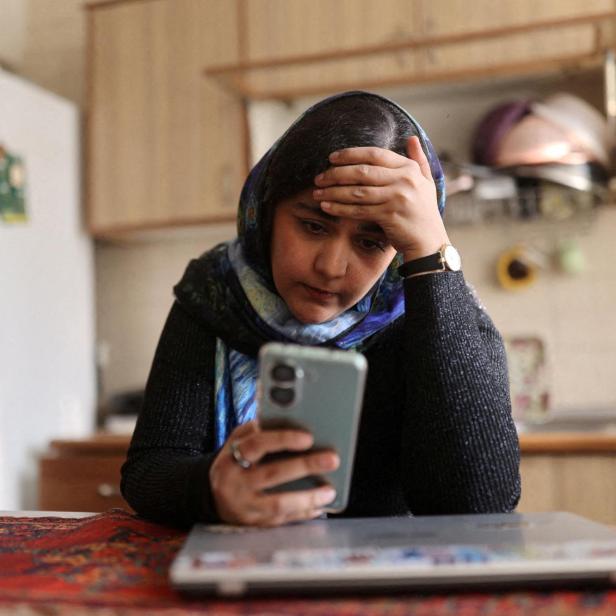 Eine Frau mit Kopftuch sitzt nachdenklich am Tisch, hält ein Smartphone in der Hand und stützt den Kopf ab.