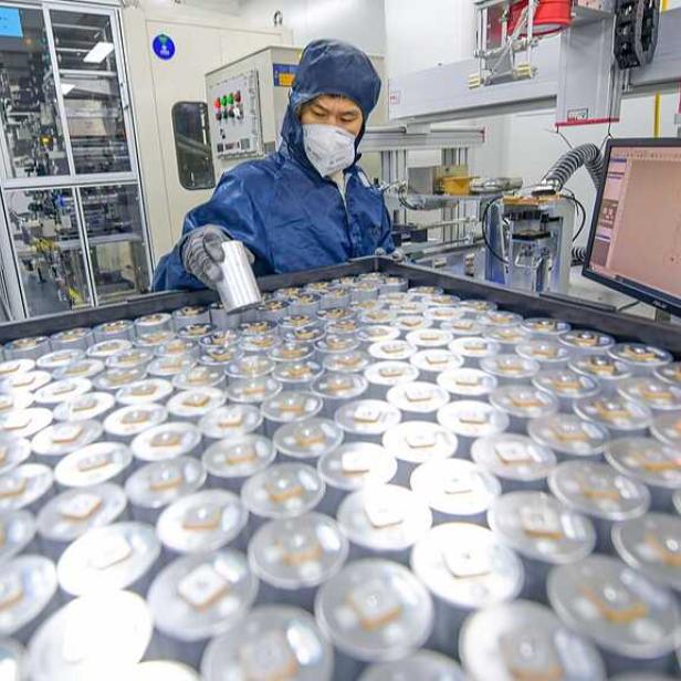 Eine Person in Schutzkleidung überprüft zahlreiche zylindrische Batteriezellen in einer modernen Fabrikumgebung.
