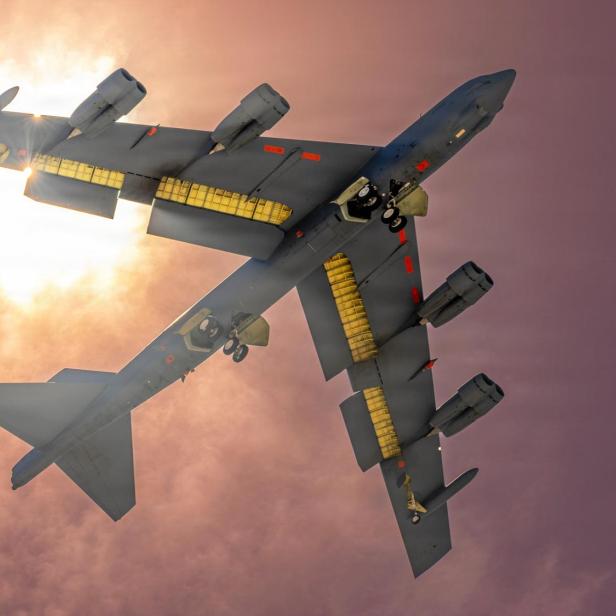 Ein B-52-Bomber fliegt mit ausgefahrenem Fahrwerk vor der Sonne durch bewölkten Himmel.