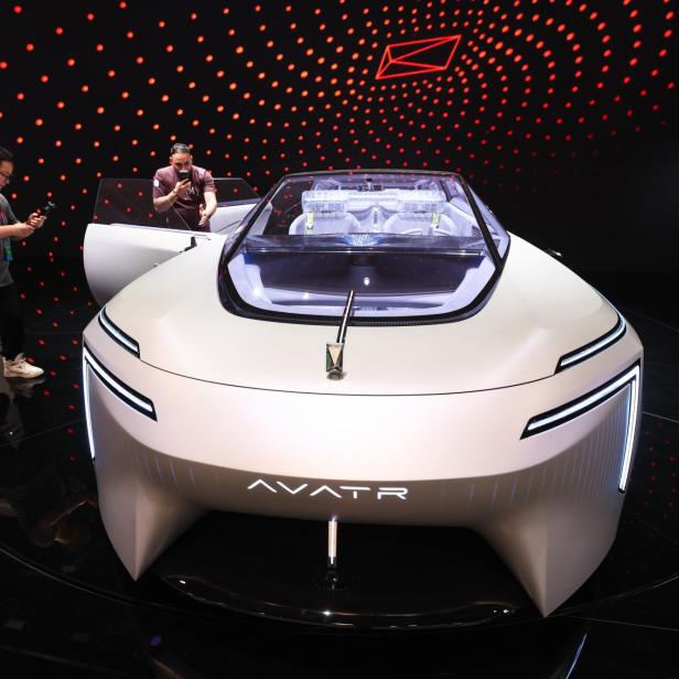 Futuristisches, weißes Konzeptauto mit offenem Dach und leuchtenden Linien, umgeben von Menschen und rotem Punktmuster im Hintergrund.