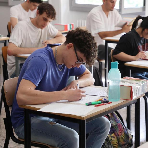 Mehrere Jugendliche sitzen an Einzeltischen in einem Klassenraum und schreiben konzentriert an Prüfungsunterlagen.