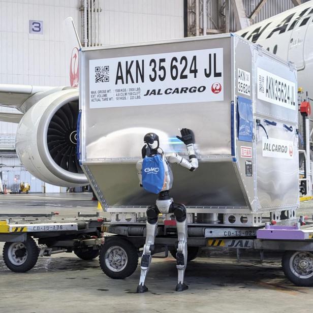 Ein humanoider Roboter mit blauem Rucksack bewegt einen großen Frachtcontainer neben einem Flugzeug in einer Lagerhalle.