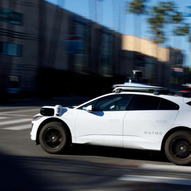 Ein weißes autonomes Auto mit Sensoren auf dem Dach fährt durch eine städtische Straße bei Tageslicht.