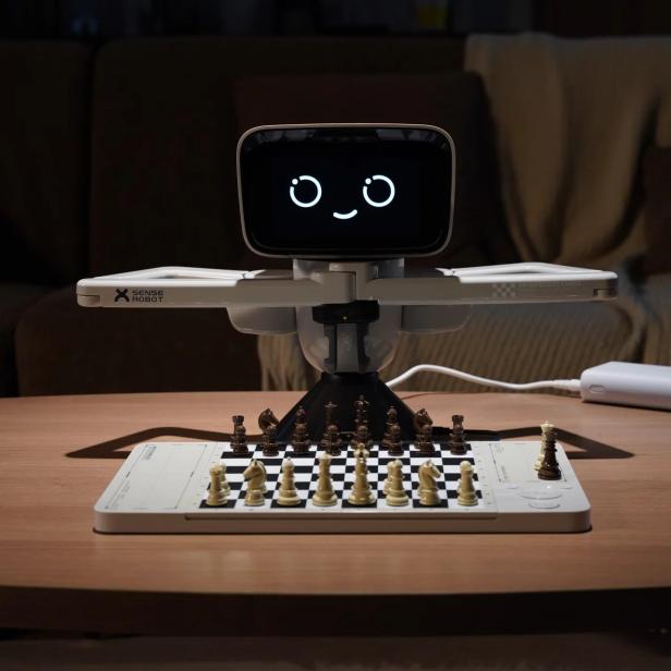 Ein Schachroboter mit Bildschirmkopf und Greifarmen steht vor einem Schachbrett auf einem Tisch.