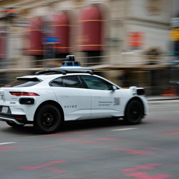 Ein weißes autonomes Auto mit Sensoren auf dem Dach fährt neben einem Polizeimotorrad durch eine Stadtstraße.