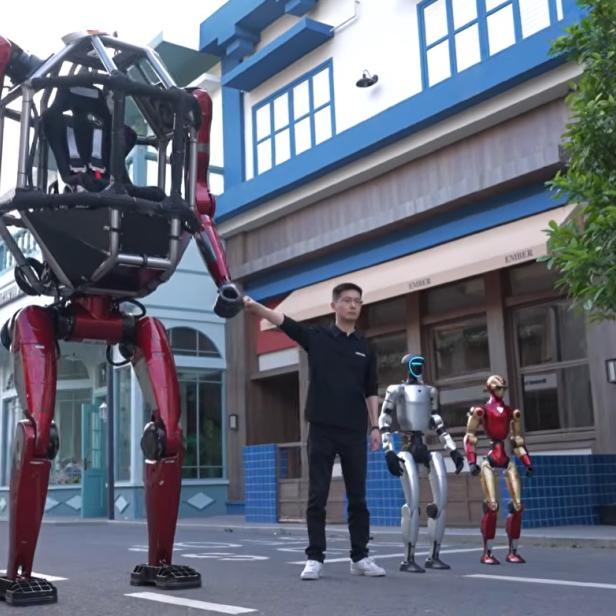 Unitree-CEO Xingxing Wang steht neben dem neuen, rot lackierten Mecha GD01 und zwei kleineren humanoiden Robotern auf einer Straße.
